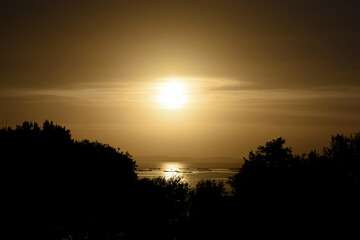 Fototapeta na wymiar Cálida y dorada puesta de sol sobre el bosque y el horizonte de la isla de Ons (Ría de Pontevedra - España). Siluetas de mejilloneras gallegas sobre el mar brillante y dorado.