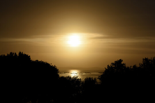 Cálida y dorada puesta de sol sobre el bosque y el horizonte de la isla de Ons (Ría de Pontevedra - España). Siluetas de mejilloneras gallegas sobre el mar brillante y dorado.
