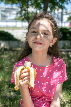 Portrait of little cute smiling girl eating banana with nice smile on sunny spring day.