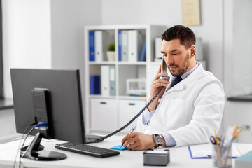 healthcare, medicine and people concept - male doctor with clipboard calling on desk phone at hospital