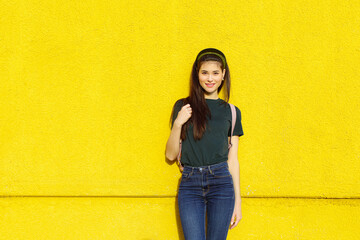Young beautiful brunette girl in jeans, a green T-shirt, hair bands and a pink backpack posing on a yellow concrete wall