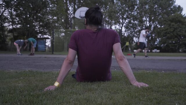 Young man sitting on the grass while resting and watching friends playing street basketball
