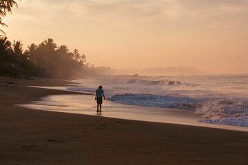 Beautiful postcard landscape shot of a woman walking on Pitiwella beach (Sri Lanka) at sunrise with orange sand, green palm trees, rocks, Indian ocean with white waves and the sun on the horizon