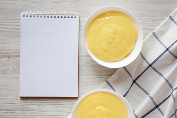 Homemade vanilla custard pudding in white bowls, blank notepad, top view. Flat lay, overhead, from above. Space for text.
