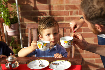 Ritratto di un bambino chiaro che fa merenda con il tè e i biscotti nel terrazzo di casa aiutato dal papà