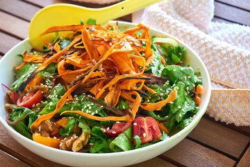 Fresh homemade salad dish in a white bowl and big yellow plastic spoon on a wooden brown table in the garden, vegan vegetable salad with cherry tomatoes, green lettuce, yam / sweet potato and walnuts