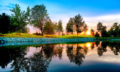 summer sunset at park panorama landscape with picturesque trees and colorful sky reflection on pond water against setting sun background. Wide view of nature in russia. Mellow evening