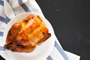 Homemade traditional rotisserie chicken on a white plate on a black surface, top view. Flat lay, overhead, from above. Copy space.