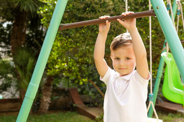 Fototapeta premium Bambino biondo con polo bianca si diverte vicino ad un altalena dentro un parco verde
