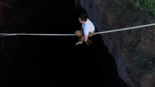 Sportsman is sitting and standing up on a slackline over the massive pit, 4k