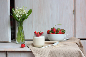 A glass jar of home made yoghurt decorated with strawberries and mint on natural color fabric. Country white wooden sideboard, a door is open. Tiny forest flowers in a glasse vase, more berries.