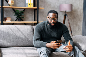 Serious African-American guy in glasses with airpods earphones sits at sofa and using smartphone for business video call. Remote work concept