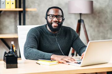 Cheerful African-American guy uses a handsfree headset and laptop to talk online at his workplace,...