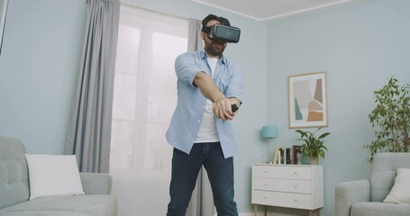 Joyful young man wearing virtual reality headset and holding controllers playing in a video game at home. Man playing VR tennis in the middle of living room. - Powered by Adobe