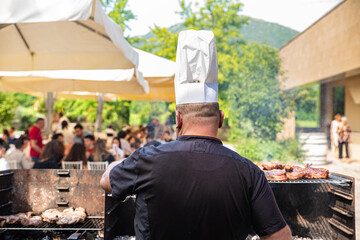 Italy. Kitchen chief with chef's hat taken from behind while cooking the meat in the barbeque. Outdoor restaurant.