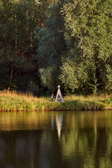 Fototapeta premium Pond on a sunny day. Wigwam on the other shore. Lake in the forest. Green fairy tale. High quality photo