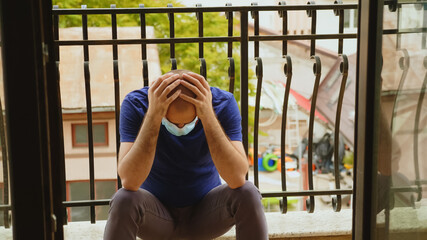 Depressed man with disposable mask sitting down in balcony during coronavirus epidemic.
