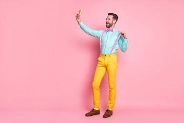 Full length photo of handsome guy trend clothes browsing telephone recording video for blog post wear shirt suspenders bow tie yellow trousers shoes isolated pastel pink color background