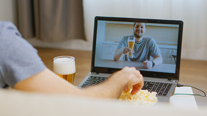 Back view of happy man on video call with his friend drinking beer during coronavirus isolation.
