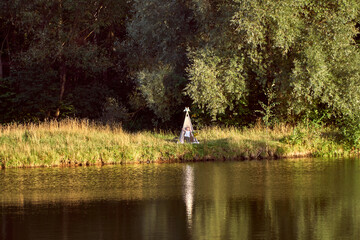 Fototapeta premium Pond on a sunny day. Wigwam on the other shore. Lake in the forest. Green fairy tale. High quality photo