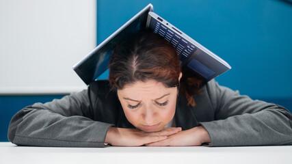 Problems at work. An exhausted woman folded her laptop over her head at her desk. Businesswoman in frustration buried under her computer. Reduction and unemployment.