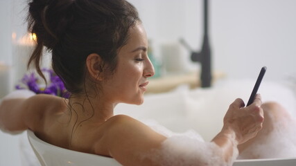 Sexy woman typing message on smartphone in bathtub. Girl lying bath with mobile