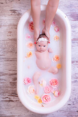 Happy kid girl takes a milk bath with petals. Bouquets of pastel rose flowers. Infant bathing. Hygiene and care for young children.