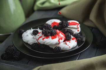 Still life, marshmallows and meringues on a black plate decorated with red berries and syrup. On a dark wooden table. The folds of the fabric on the dark boards.