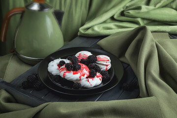 White meringue and marshmallows on a black plate, poured with red syrup. Strawberry syrup. Dessert is decorated with berries. Black berries on white cookies. Still life on a dark wooden table.