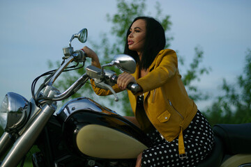 A woman in a black dress and yellow jacket sits on a motorcycle against the background of trees.