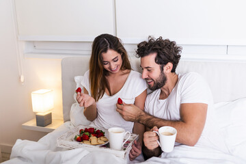 Obraz na płótnie Canvas Cute couple having breakfast in bed in the bedroom. Beautiful woman feeding her boyfriend strawberries in bed while having breakfast and coffee in bedroom