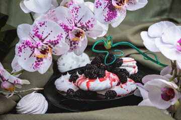 Obraz na płótnie Canvas White meringue and marshmallows on a black plate, poured with red syrup. Still life of flowers and baking. Light Orchid. Strawberry syrup. Dessert is decorated with berries. 