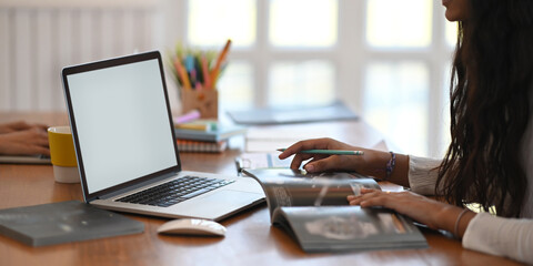 A young designer is working with a white blank screen computer laptop at the wooden working desk.