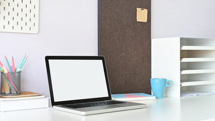 A white blank screen computer laptop is putting on a white working desk surrounded by office equipment.