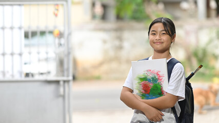 A schoolgirl is holding painting equipment and carrying a school bag while standing and waiting for a school bus.