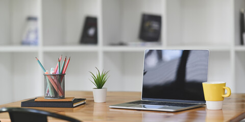 A black empty screen laptop is putting on a wooden working desk surrounded by office equipment.