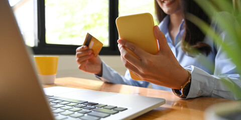 A beautiful woman is paying bills via mobile phone and credit card at the wooden working desk.