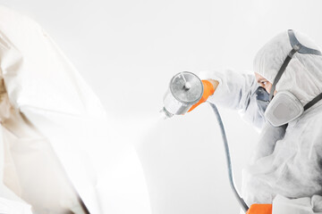 Body painter with airgun spraying white paint on a automobile in car maintenance service paint room.