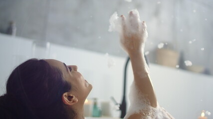 Close up relaxed woman blowing foam in bathroom. Happy girl having fun with foam