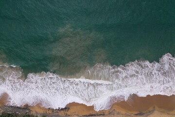Naklejka na ściany i meble aerial drone bird view shot of the sea shore with yellow sand, black rocks, large white waves and foam crashing on the beach forming beautiful textures, patterns, shapes. Sri Lanka
