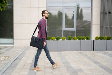 Attractive bearded man in eyeglasses walking on city street. Guy with laptop in bag going somewhere on pavement. Lifestyle and leisure concept