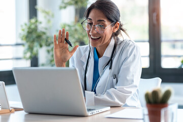 Female doctor waving and talking with colleagues through a video call with a laptop in the...