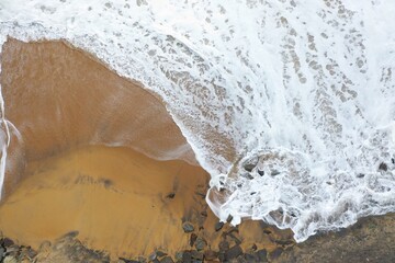 aerial drone bird view shot of the sea shore with yellow sand, black rocks, large white waves and foam crashing on the beach forming beautiful textures, patterns, shapes. Sri Lanka