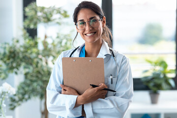 Beautiful female doctor looking at camera and smiling while holding medical documents and standing...