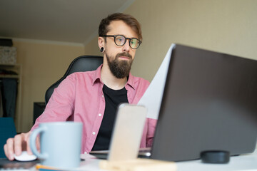 Bearded young man in eyeglasses working at home. Dark-haired attractive guy looking at laptop screen and sitting on isolation. Workplace and freelance concept