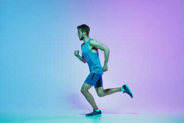 Fototapeta na wymiar Portrait of active young caucasian man running, jogging on gradient studio background in neon light. Professional sportsman training in action and motion. Sport, wellness, activity, vitality concept.