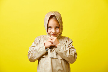 Caucasian little girl in wet demi-season jacket with hood standing on colored yellow background.