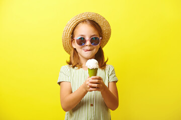 Beautiful little girl holding an ice cream and going to eat it, licking her lips, dressed in a summer dress, straw hat and sunglasses, standing on yellow isolated.