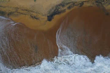 aerial drone bird view shot of the sea shore with yellow sand, black rocks, large white waves and foam crashing on the beach forming beautiful textures, patterns, shapes. Sri Lanka