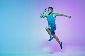 Fototapeta na wymiar In jump. Portrait of young caucasian man running, jogging on gradient studio background in neon light. Professional sportsman training in action and motion. Sport, wellness, activity, vitality concept